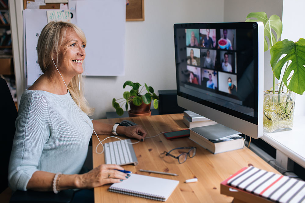 An older woman smiles at a computer screen, which shows the faces of other participants of a virtual meeting.