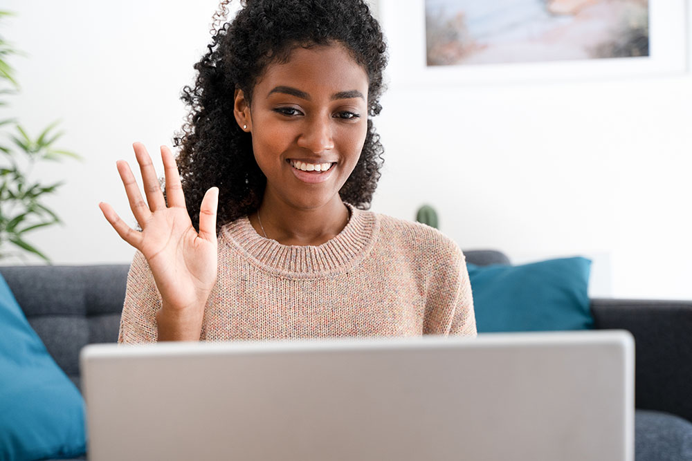 A Black woman behind a computer screen waves hello to fellow paticipants over a virtual meeting.