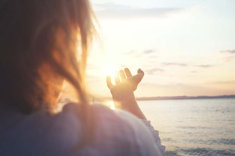 A woman extends her hand toward the ocean as the sun sets in the background.