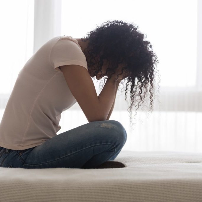 Upset sad African American woman suffering from loneliness side view, sitting in bed alone, holding head in hands, upset girl has psychological troubles, trauma, thinking about problem in bedroom