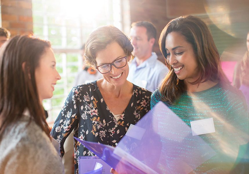 Three women with nametags look down at a conference program together.