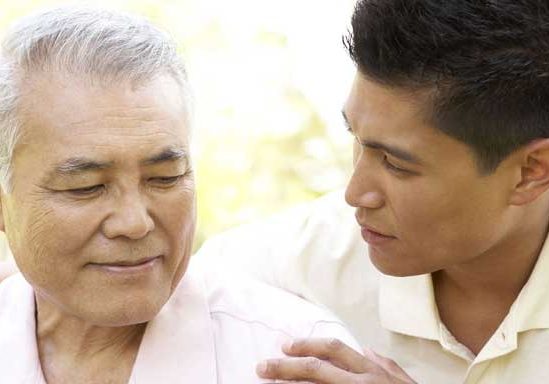 A young man places his hands on the shoulders of another older man, helping him.